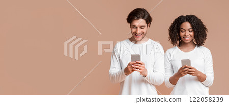 Gadget addiction. Smiling interracial couple busy with their smartphones, looking at device screen and ignoring each other, yellow background Gadget addiction. Smiling interracial couple busy with their smartphones, looking at device screen and ignoring each other, yellow background 122058239
