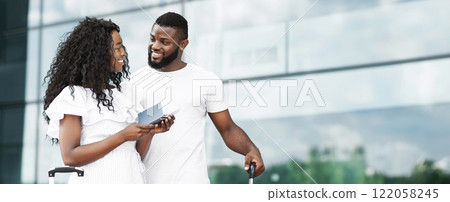 Happy African American couple standing together at the airport. The woman is wearing a white dress and has long curly hair. They are both smiling and looking at each other, copy space 122058245
