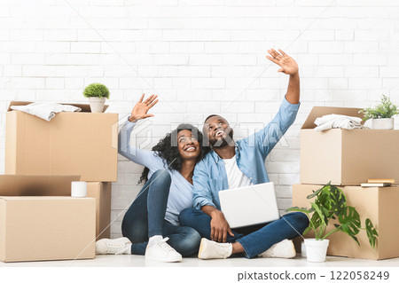 New house decorating. Young african couple sitting on floor at new home and pointing at copy space, using laptop 122058249