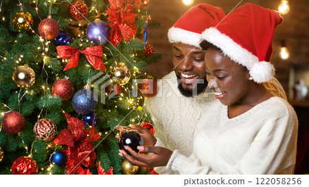 Happy holidays. African american couple in Santa hats decorating Christmas tree together Happy holidays. African american couple in Santa hats decorating Christmas tree together 122058256