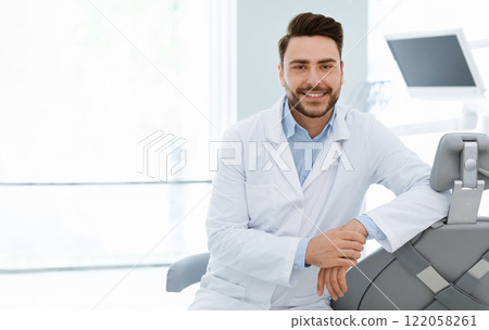 Attractive young bearded man dentist keeping beautiful smile on his face, posing over modern dental cabinet background, empty space. People occupations, healthcare, dentistry concept 122058261