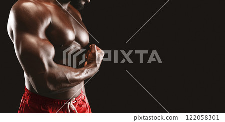 A muscular man in red shorts flexes his bicep against a black background. He is showcasing his strength and fitness. 122058301
