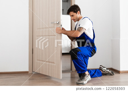 Installation of a lock on the front wooden entrance door. Portrait of young locksmith workman in blue uniform installing door knob. Professional repair service. Maintenance Concept 122058303