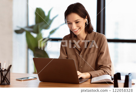 Happy Worker. Portrait of smiling girl in headphones using laptop, sitting at office and listening to music, copyspace 122058328