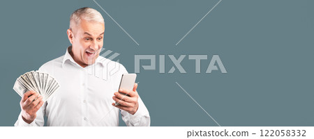 Cashback Concept. Surprised senior man holding bunch of money banknotes, using smartphone, copyspace, blue studio wall 122058332