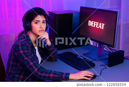 A young woman, wearing headphones, sits at a computer desk with a serious expression on her face. The word DEFEAT is displayed on the computer screen, indicating that she has lost a game 122058354