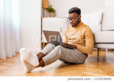 Shocked african american man using computer reading news on laptop sitting on floor at home. Selective focus Shocked african american man using computer reading news on laptop sitting on floor at home. Selective focus 122058400