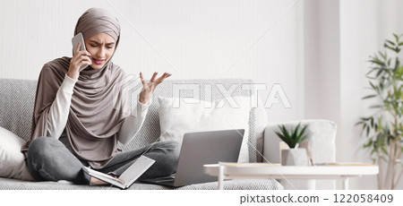 Freelance stress. Concerned muslim girl in hijab talking on cellphone and emotionally gesturing while working online on laptop at home, free space Freelance stress. Concerned muslim girl in hijab talking on cellphone and emotionally gesturing while working online on laptop at home, free space 122058409