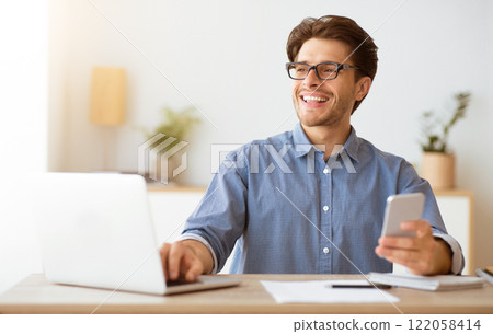 A man wearing glasses sits at a desk working on his laptop, smiling as he holds a phone in his other hand. A man wearing glasses sits at a desk working on his laptop, smiling as he holds a phone in his other hand. 122058414
