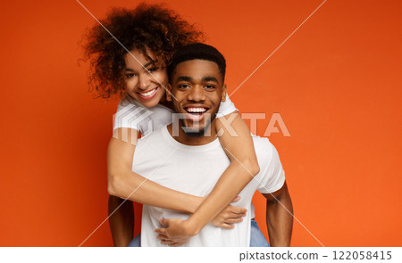 Man giving piggyback ride to his girlfriend. African-american couple having fun on orange background 122058415