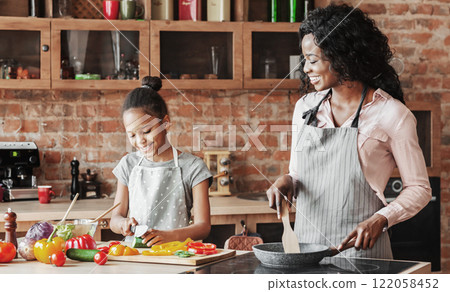 Little sweet african american girl helping her mom at kitchen, making healthy dinner, copy space Little sweet african american girl helping her mom at kitchen, making healthy dinner, copy space 122058452
