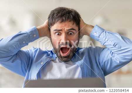 A young man sits at his computer, looking shocked and holding his head in his hands as he stares at the screen. He is likely reacting to something he saw on the laptop 122058454