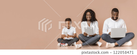 Modern family. Happy african american father, mother and daughter using different gadgets while sitting on floor over yellow studio background, panorama 122058471