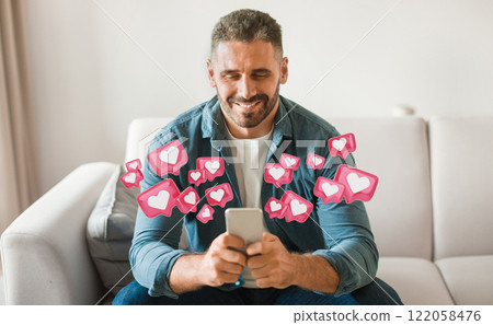 A man is sitting on a white couch, holding a smartphone in his hands, checking his social media. He is smiling as he looks down at his phone. Surrounding him are many pink like icons. 122058476