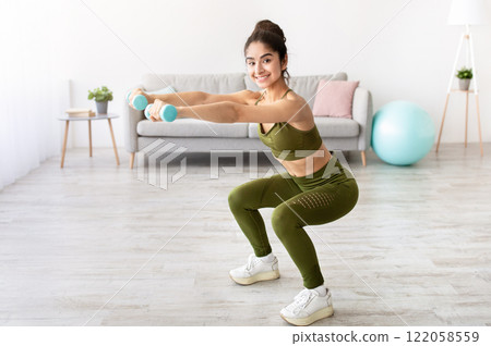 Strong young Indian woman working out with dumbbells, squatting at home, empty space. Fit Eastern lady taking care of her health, having domestic training, leading active lifesyle 122058559