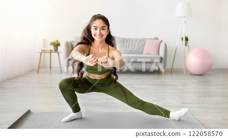 Lockdown home workout. Sporty young Indian lady stretching legs on yoga mat in living room, free space. Positive Eastern woman doing flexibility exercises, keeping fit and healthy, enjoying training Lockdown home workout. Sporty young Indian lady stretching legs on yoga mat in living room, free space. Positive Eastern woman doing flexibility exercises, keeping fit and healthy, enjoying training 122058570