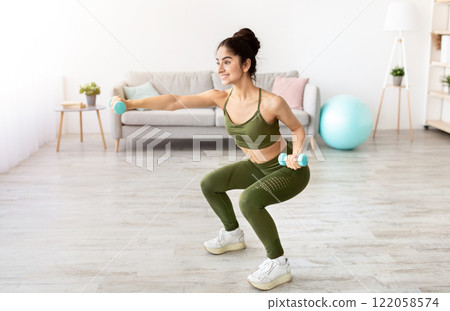 Happy young Indian woman doing squats with dumbbells at home, full length. Positive millennial Asian lady performing strength exercises, keeping in good shape during covid quarantine 122058574