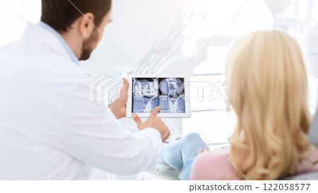 Man dentist showing female patient treatment goal, using digital tablet, dental clinic interior. Bearded man stomatologist having conversation with lady in dental chair, pointing at gadget, back view 122058577