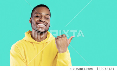 Smiling African Guy Pointing Thumb Finger Aside At Blank Space For Text Advertising Your Offer Standing Over Blue Background, Studio Shot. Look There Concept. Panorama 122058584