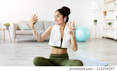 Joyful Indian woman taking selfie on break from home workout, full length. Happy Asian lady with smartphone making photo of herself on domestic training, making peace gesture and smiling at webcam 122058597