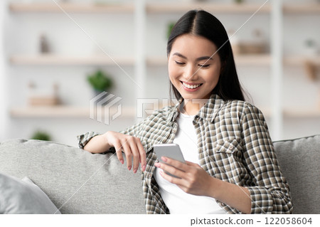 Cheerful asian lady sitting on sofa and using smartphone, chatting with girlfriend or lover, copy space. Pretty young woman with mobile phone chilling on couch, checking social media, home interior 122058604
