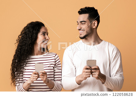 Mobile Communication. Cheerful Arab Man And Woman Holding Smartphones And Looking At Each Other, Happy Middle Eastern Couple Using Cellphones While Standing Together On Yellow Background, Copy Space 122058628