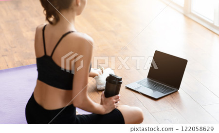Rear back over the shoulder view of unrecognizable woman sitting on yoga mat drinking water and looking at blank laptop screen, having video call with trainer or attending fitness class online, mockup 122058629