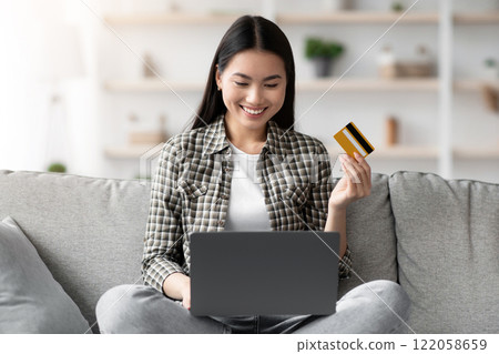 Cheerful young asian woman sitting on couch in living room and using laptop and credit card, checking new webstore for online shopping or banking, home interior, copy space, catching sales Cheerful young asian woman sitting on couch in living room and using laptop and credit card, checking new webstore for online shopping or banking, home interior, copy space, catching sales 122058659
