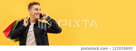 Positive handsome young guy wearing casual clothing with colorful shopping bags on his shoulder talking on phone and looking at copy space for advertisement, yellow background, web-banner 122058693