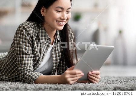 Positive asian woman surfing on social media, using digital tablet, living room interior, copy space. Happy young korean lady laying on floor and reading blog on modern pad, enjoying new mobile app 122058701