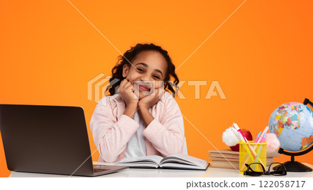 Portrait Of Cute And Shy Black Teenage Girl Touching Cheeks With Fists And Smiling At Camera, Young Curly Afro Lady Posing Sitting At Desk With Pc Isolated Over Yellow Studio Background, Banner 122058717