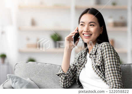 Cheerful asian lady sitting on sofa and talking on smartphone with girlfriend or lover, looking at copy space and smiling. Pretty young woman having phone conversation, home interior 122058822