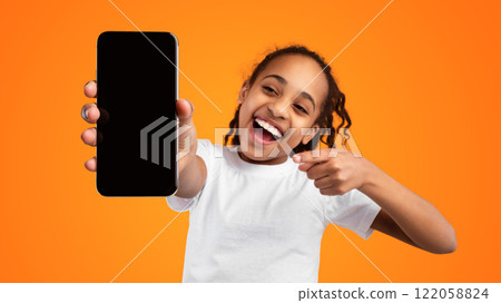 Trendy Mobile Phone. Excited young black girl holding smartphone with empty screen in hand, showing device close to camera and pointing at gadget with blank space, mockup, yellow studio background Trendy Mobile Phone. Excited young black girl holding smartphone with empty screen in hand, showing device close to camera and pointing at gadget with blank space, mockup, yellow studio background 122058824