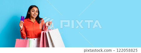 Shopping. Happy Young Indian Woman Shopper With Paper Bags In Hands Happily Showing Her Credit Card, Showcasing Excitement Over Sale Offer On Blue Background, Studio Shot 122058825
