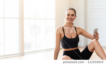 Well fit athletic lady with wireless headphones and smartphone having break, sitting on fitness mat doing home workout, listening to favorite music playlist, looking away and smiling, free copy space 122058934