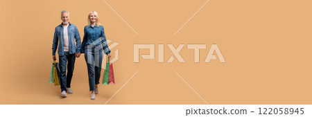 Retail, shopping, season sale. Positive happy beautiful retired senior man and woman wearing casual denim outfit with shopping bags holding hands, walk over beige background, full length, copy space 122058945