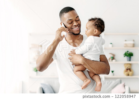 Multitasking Concept. Young Black Man Holding Little Baby And Talking On Cellphone While Working Remotely With Laptop At Home, African American Freelancer Guy Enjoying Paternity Leave, Copy Space 122058947