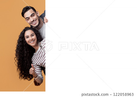 Happy Arabic Man And Woman Peeking Out Behind Big White Advertisement Board, Cheerful Smiling Middle Eastern Couple Demonstrating Free Copy Space On Blank Billboard, Posing Over Yellow Background 122058963