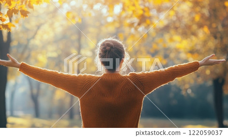 A woman stands in a park with her arms outstretched, smiling as she enjoys the bright sun shining through the golden leaves of the trees. 122059037
