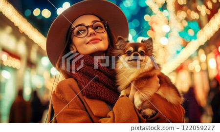A young woman wearing a fashionable hat smiles gently while holding a small dog close to her. The background features colorful lights and decorations, creating a joyful atmosphere. 122059227