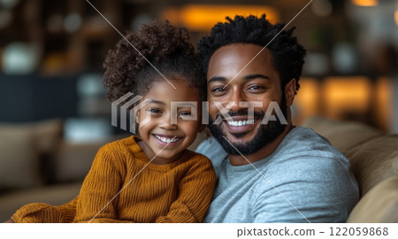 A father and his young daughter smile widely while sitting closely together on a sofa in their home, radiating happiness and warmth during a peaceful afternoon. 122059868