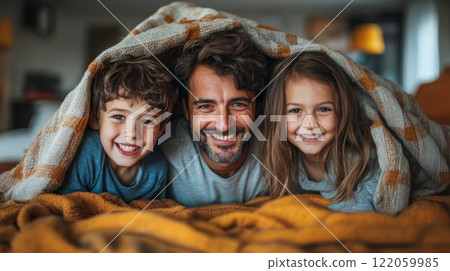 A father enjoys a joyful morning indoors with his two children, all snuggled happily under a cozy blanket in a colorful, inviting space. 122059985