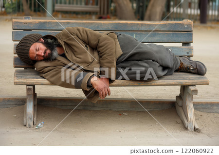 Poor homeless man or refugee sleeping on the wooden bench on the urban street in the city 122060922