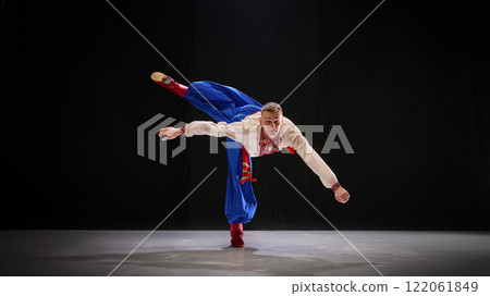 Dancer in white embroidered shirt and blue trousers balances gracefully on one foot, leaning forward into dynamic pose, evoking agility of Ukrainian folk traditions 122061849