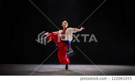 Male dancer in folk-style outfit performs elegant leap, arms extended, radiating confidence and joy, showcasing celebratory nature of Hopak. 122061855