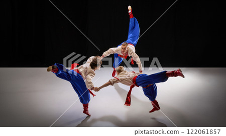 Three dancers hold hands mid-performance, creating motion with high kicks and synchronized movements, symbolizing unity in Hopak. Three dancers hold hands mid-performance, creating motion with high kicks and synchronized movements, symbolizing unity in Hopak. 122061857