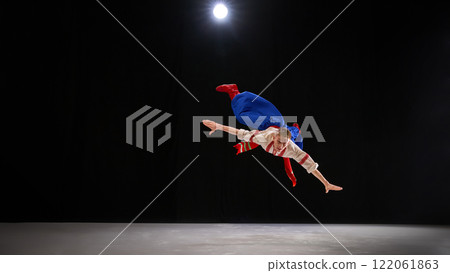 Performer dynamically leaping with arms and legs extended, illustrating energetic Hopak dance movement under focused stage spotlight on black backdrop. 122061863