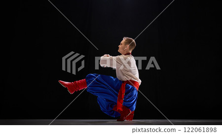 Male dancer in deep squat with one leg extended, arms elegantly balanced, showcasing agility and tradition against black studio background. 122061898