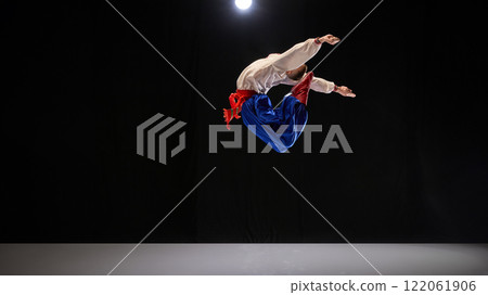 Male dancer executing backward jump, dressed in traditional embroidered shirt, blue trousers, and red boots, expressing culture and movement against black studio background. Male dancer executing backward jump, dressed in traditional embroidered shirt, blue trousers, and red boots, expressing culture and movement against black studio background. 122061906
