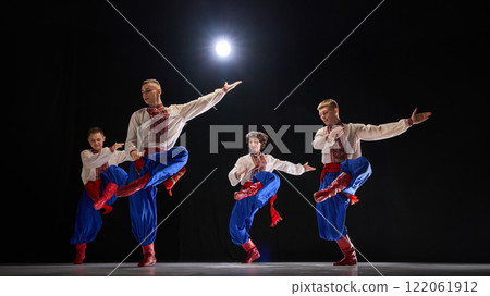 Male dancer leaping with arms stretched, traditional attire flowing, showcasing rhythmic motion and cultural storytelling against black studio background. 122061912